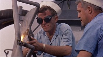 Movie still from “Operation Petticoat” (1959), directed by Blake Edwards – A man wearing a hat and glasses welding a pipe; Medium shot, Low angle