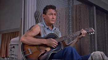 Movie still from “Operation Petticoat” (1959), directed by Blake Edwards – A man holding a guitar while sitting on a chair; Medium shot, High angle