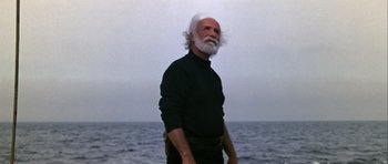 Movie still from “Orca” (1977), directed by Michael Anderson – An older man with a white beard standing in front of a body of water; Medium shot, Low angle
