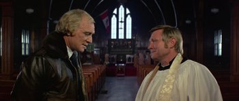 Movie still from “Orca” (1977), directed by Michael Anderson – Two men standing in front of each other in front of a church; Medium shot, Low angle