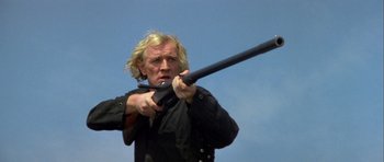 Movie still from “Orca” (1977), directed by Michael Anderson – A man holding a rifle in front of a blue sky; Close Up shot, Low angle
