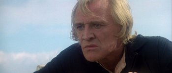 Movie still from “Orca” (1977), directed by Michael Anderson – A man with long blonde hair and wearing a black shirt; Close Up shot, Low angle