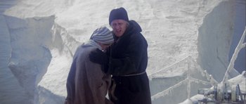Movie still from “Orca” (1977), directed by Michael Anderson – Two people in winter coats and hats are standing in the snow; Medium shot, High angle