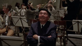Movie still from “Orchestra Rehearsal” (1978), directed by Federico Fellini – An older man sitting in a chair in front of a group of men playing brass instruments; Medium shot, Low angle