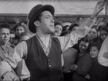 Movie still from “Obsession” (1943), directed by Luchino Visconti – A man wearing a vest and a hat in front of a group of people; Medium shot, Low angle