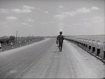 Movie still from “Obsession” (1943), directed by Luchino Visconti – A man walking down the middle of a road holding a surfboard; Wide shot, High angle