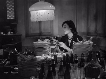 Movie still from “Obsession” (1943), directed by Luchino Visconti – A woman in a black jacket is setting plates on a table; Medium shot, High angle