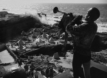 Movie still from “Othello” (1951), directed by Orson Welles – A man playing a trumpet on the beach; Extreme Wide shot, High angle