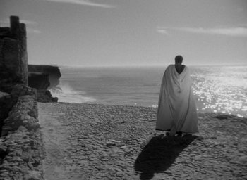 Movie still from “Othello” (1951), directed by Orson Welles – A man standing on a beach with a blanket over his head; Extreme Wide shot, High angle