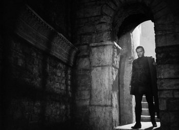 Movie still from “Othello” (1951), directed by Orson Welles – A man standing in an archway in an old building; Wide shot, Low angle