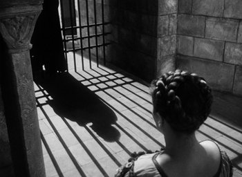 Movie still from “Othello” (1951), directed by Orson Welles – A woman sitting in front of a metal gate; Wide shot, Low angle