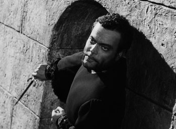 Movie still from “Othello” (1951), directed by Orson Welles – Black and white photograph of a man holding a sword in front of a stone wall; Medium shot, High angle