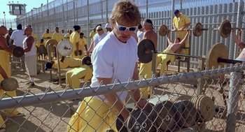 Movie still from “Out of Sight” (1998), directed by Steven Soderbergh – A man in white shirt and yellow pants holding a barbell; Wide shot, Over the shoulder angle