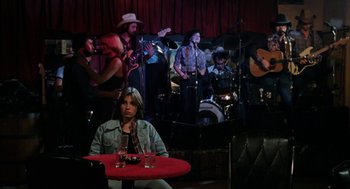 Movie still from “Out of the Blue” (1980), directed by Dennis Hopper – A woman sitting at a table in front of a group of people; Wide shot, Over the shoulder angle