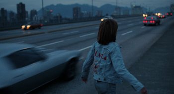 Movie still from “Out of the Blue” (1980), directed by Dennis Hopper – A woman is walking down the street with a car behind her; Wide shot, Over the shoulder angle