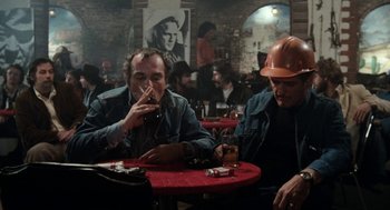 Movie still from “Out of the Blue” (1980), directed by Dennis Hopper – Two men sitting at a table in front of a wall; Medium shot, Over the shoulder angle