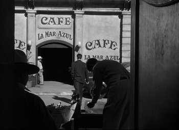 Movie still from “Out of the Past” (1947), directed by Jacques Tourneur – A man standing in front of a building with people standing around; Wide shot, Over the shoulder angle