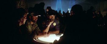 Movie still from “Outland” (1981), directed by Peter Hyams – A group of men sitting at a table in a dark room; Medium shot, Over the shoulder angle