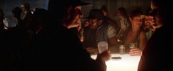 Movie still from “Outland” (1981), directed by Peter Hyams – A group of people sitting at a table with drinks; Medium shot, Over the shoulder angle