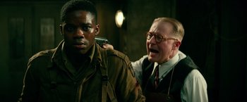 Movie still from “Overlord” (2018), directed by Julius Avery – An older man with glasses is holding an older man with a gun; Medium shot, Over the shoulder angle
