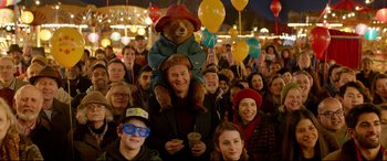 Movie still from “Paddington 2” (2017), directed by Paul King – A crowd of people with balloons and a teddy bear on their shoulders; Medium shot, Low angle