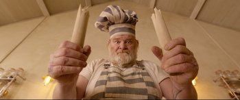 Movie still from “Paddington 2” (2017), directed by Paul King – An old man wearing a chef's hat and apron holding a pair of wooden utensils in front of his face; Close Up shot, Low angle