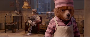 Movie still from “Paddington 2” (2017), directed by Paul King – A man in a prison uniform reading a newspaper while a woman in a pink apron looks on; Medium shot, Low angle