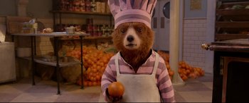 Movie still from “Paddington 2” (2017), directed by Paul King – A bear dressed as a chef holding an orange in a grocery store; Medium shot, High angle
