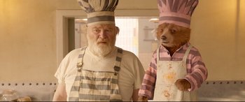 Movie still from “Paddington 2” (2017), directed by Paul King – A man and a dog wearing aprons standing next to each other in a kitchen; Close Up shot, Low angle