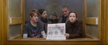 Movie still from “Paddington 2” (2017), directed by Paul King – A group of people sitting around a table holding a newspaper; Medium shot, Over the shoulder angle