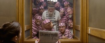 Movie still from “Paddington 2” (2017), directed by Paul King – A group of men in pink and white striped uniforms are gathered around a newspaper; Wide shot, Overhead angle