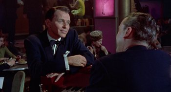 Movie still from “Pal Joey” (1957), directed by George Sidney – A man sitting at a table in front of two other people; Medium shot, Over the shoulder angle