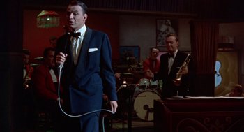 Movie still from “Pal Joey” (1957), directed by George Sidney – A man in a suit and tie holding a microphone in front of a group of people; Medium shot, Low angle