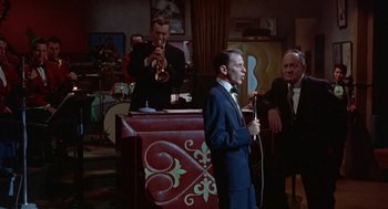 Movie still from “Pal Joey” (1957), directed by George Sidney – A man in a suit and tie holding a microphone; Medium shot, Over the shoulder angle
