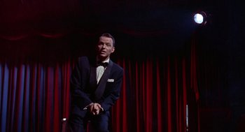 Movie still from “Pal Joey” (1957), directed by George Sidney – A man wearing a suit and bow tie sitting in front of a curtain; Medium shot, Low angle