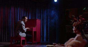 Movie still from “Pal Joey” (1957), directed by George Sidney – A man sitting in front of a piano in a dark room; Wide shot, High angle