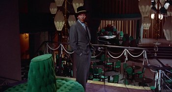 Movie still from “Pal Joey” (1957), directed by George Sidney – A man in a suit standing in front of green chairs; Wide shot, High angle