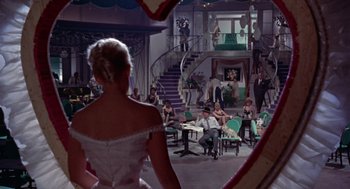 Movie still from “Pal Joey” (1957), directed by George Sidney – A woman in a white dress standing in front of a group of people in a room; Wide shot, High angle