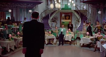 Movie still from “Pal Joey” (1957), directed by George Sidney – A man standing in front of a dining room table; Wide shot, High angle
