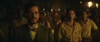 Movie still from “Palm Trees in the Snow” (2015), directed by Fernando González Molina – A man in a suit and bow tie standing in front of a group of people; Close Up shot, Over the shoulder angle