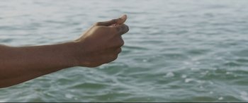 Movie still from “Palm Trees in the Snow” (2015), directed by Fernando González Molina – A hand is holding something in front of a body of water; Extreme Close Up shot, Overhead angle