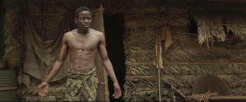 Movie still from “Palm Trees in the Snow” (2015), directed by Fernando González Molina – A man standing in front of a building with a straw hut; Medium shot, Low angle