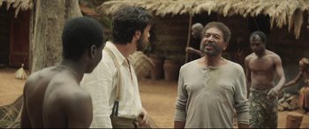 Movie still from “Palm Trees in the Snow” (2015), directed by Fernando González Molina – Two men talking to each other in front of an african hut; Medium shot, Over the shoulder angle