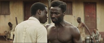 Movie still from “Palm Trees in the Snow” (2015), directed by Fernando González Molina – Two men standing next to each other in front of a building; Close Up shot, Over the shoulder angle