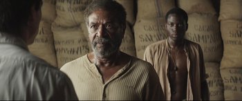 Movie still from “Palm Trees in the Snow” (2015), directed by Fernando González Molina – A man standing in front of bags of coffee; Medium shot, Over the shoulder angle
