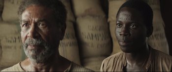 Movie still from “Palm Trees in the Snow” (2015), directed by Fernando González Molina – Two men standing next to each other in front of bags of coffee; Close Up shot, Over the shoulder angle