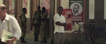 Movie still from “Palm Trees in the Snow” (2015), directed by Fernando González Molina – A man standing in front of a building next to two soldiers in fatigues; Medium shot, Over the shoulder angle
