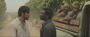 Movie still from “Palm Trees in the Snow” (2015), directed by Fernando González Molina – Two men standing next to each other on a dirt road; Medium shot, Over the shoulder angle
