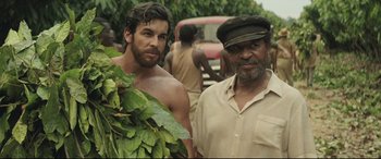 Movie still from “Palm Trees in the Snow” (2015), directed by Fernando González Molina – Two men standing next to each other near a street; Medium shot, Over the shoulder angle