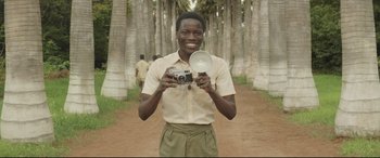 Movie still from “Palm Trees in the Snow” (2015), directed by Fernando González Molina – A man holding a camera in front of some trees; Medium shot, Low angle